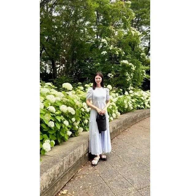 女性が白いヒナギクの花壇の前に立っている。薄い青いロングドレスを着て、黒いトートバッグを手にしている。背景には緑豊かな木々が広がっている。