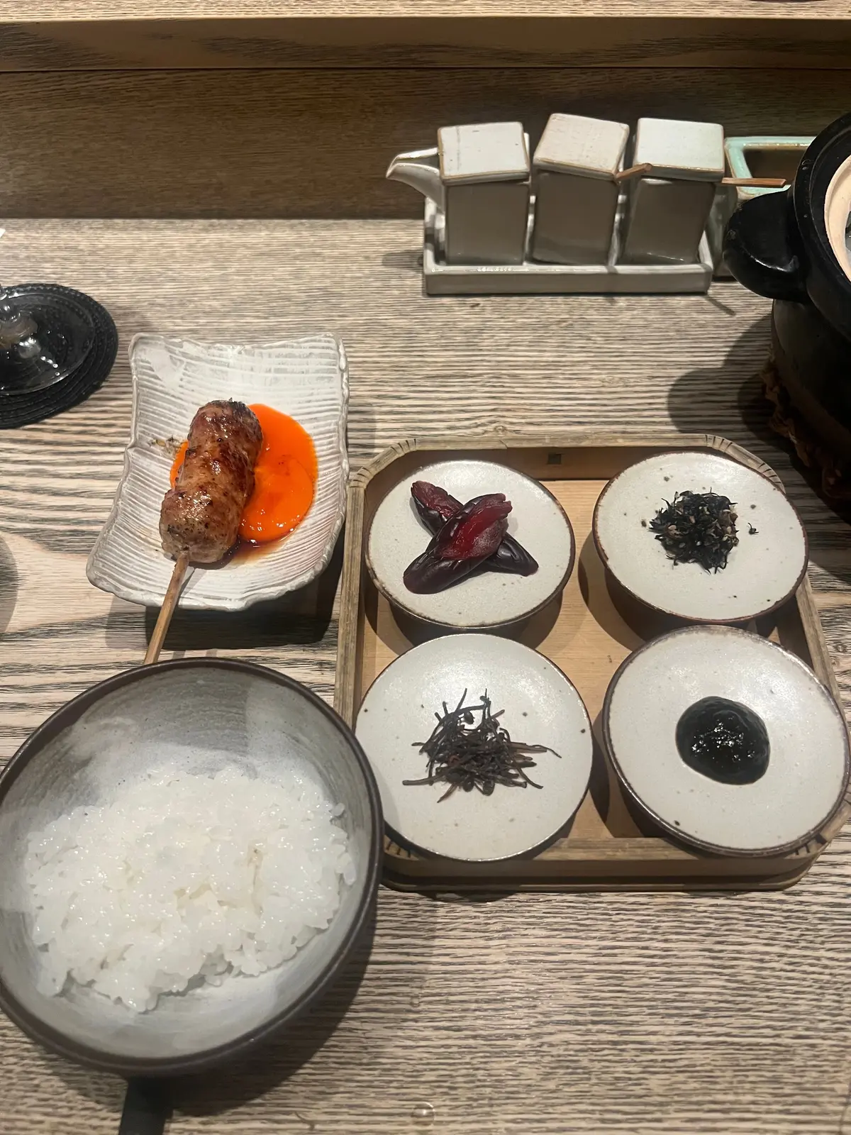 木製のトレイに、串に刺したソーセージとオレンジ色のソース、小さな白い皿に赤紫のフルーツ、黒い海藻の盛り合わせ、米飯が入ったボウルなど、精巧に盛り付けられた料理が並んでいる。