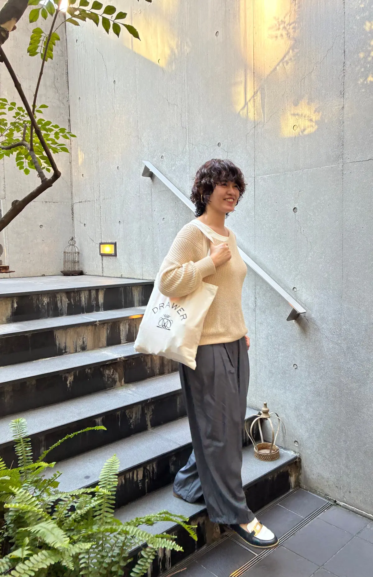 白いトートバッグを肩にかけた女性が、コンクリートの階段を背景に横を向いて微笑んでいる。茶色の巻き髪で、白いニットと黒いパンツを着用。トートバッグには「DRAWER」のロゴマークがプリントされている。