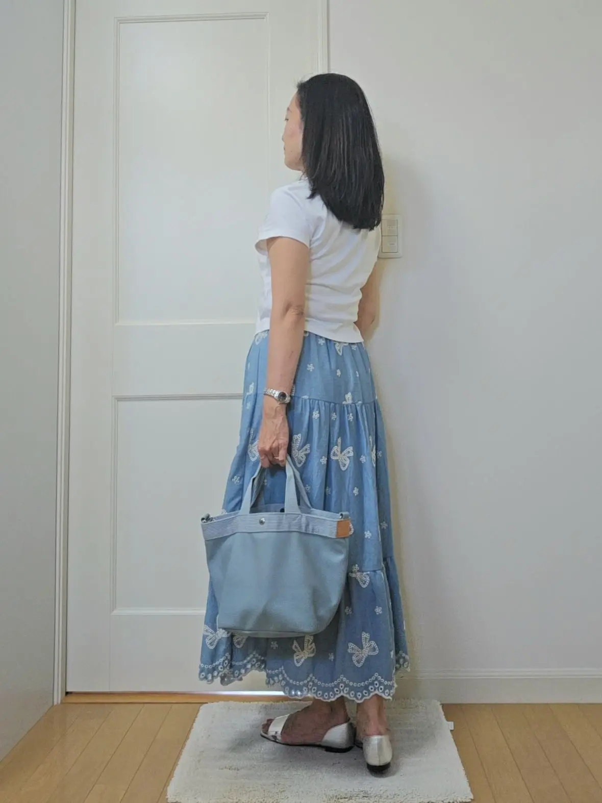 白いTシャツに青い蝶柄のティアードスカート、シルバーのサンダルを身につけた女性が、ドアの前で振り返ってこちらを見ている。左手に青いトートバッグを持っている。