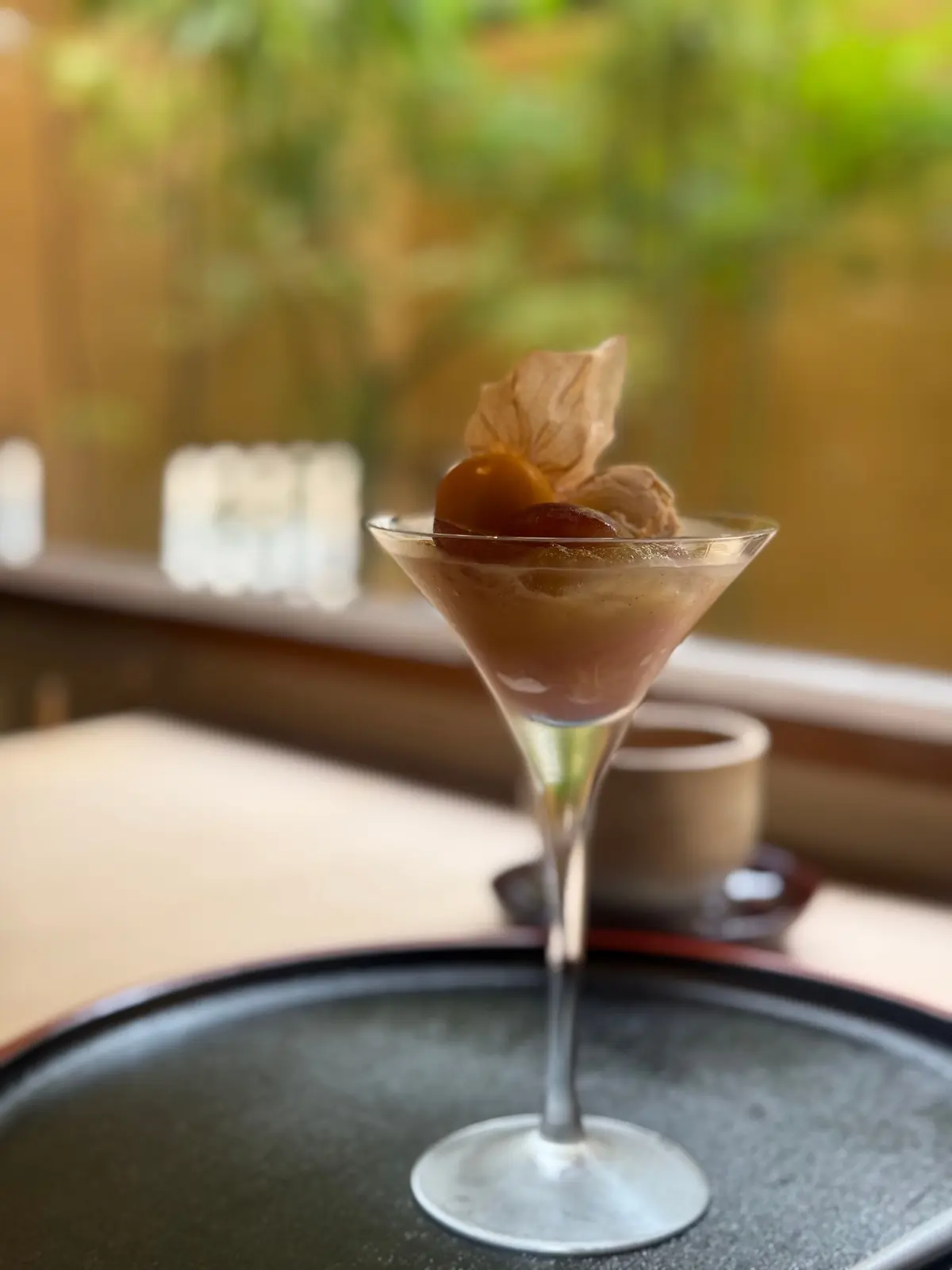 マーティニグラスに盛り付けられた、茶色のチョコレートムース。表面にはチョコレートボールと、チョコレート色の葉が飾られている。黒いお皿の上に置かれ、背景には緑の植物が見える。