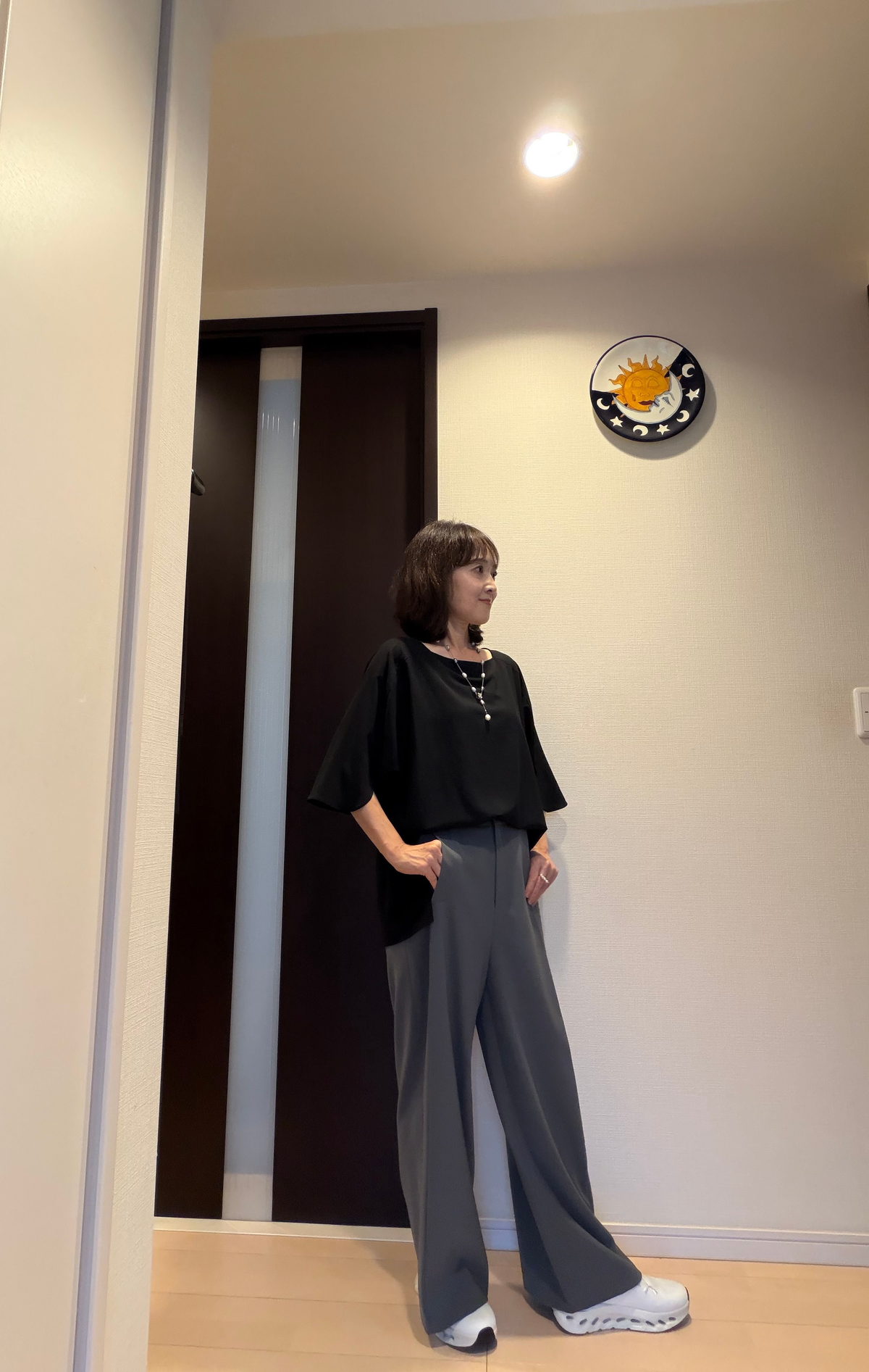 黒いトップスとグレーのワイドパンツを着用した女性が立っている全身写真。白いスニーカーを履き、短髪で微笑んでいる。