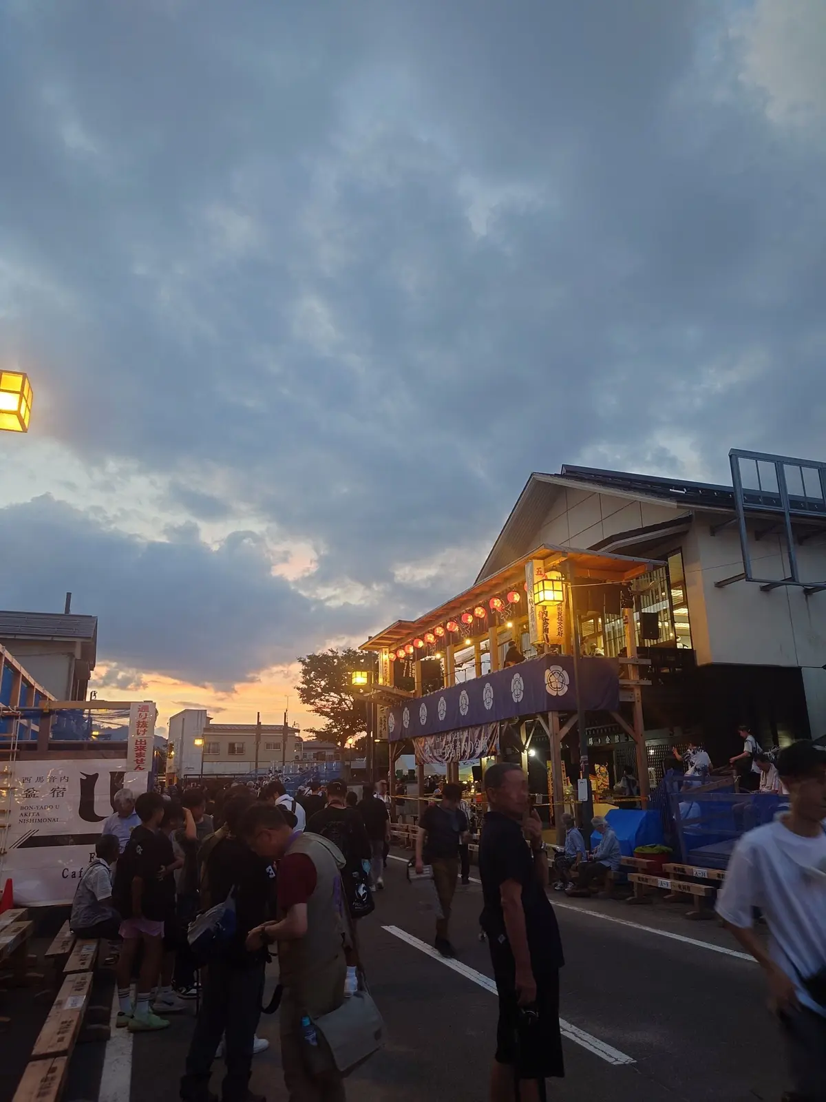 夕暮れ時の市場の様子。多くの人が集まり、屋台や店舗が並ぶ。木造の建物には提灯が灯り、活気がある。空は雲が多く、オレンジ色の光が差し込んでいる。