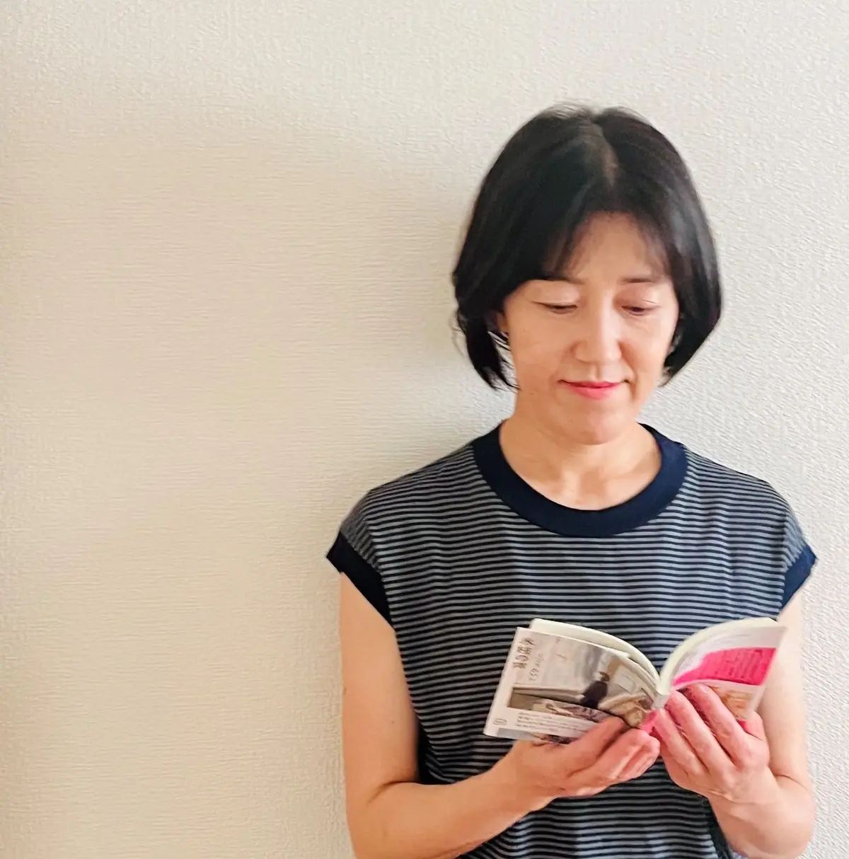 女性が白い壁に寄りかかり、雑誌を手に持って読んでいる。黒と白の縞模様の半袖シャツを着ている。