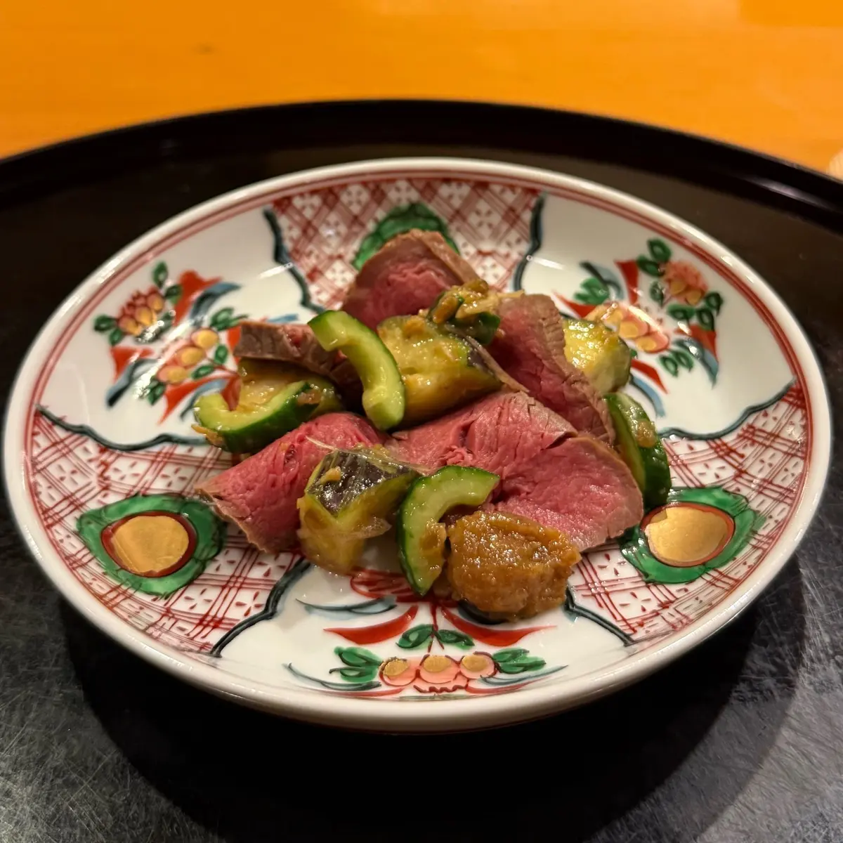 赤い肉と緑色の野菜が盛り付けられた、白地に赤と緑の模様が入ったお皿。黒い木製のトレイに乗せられている。