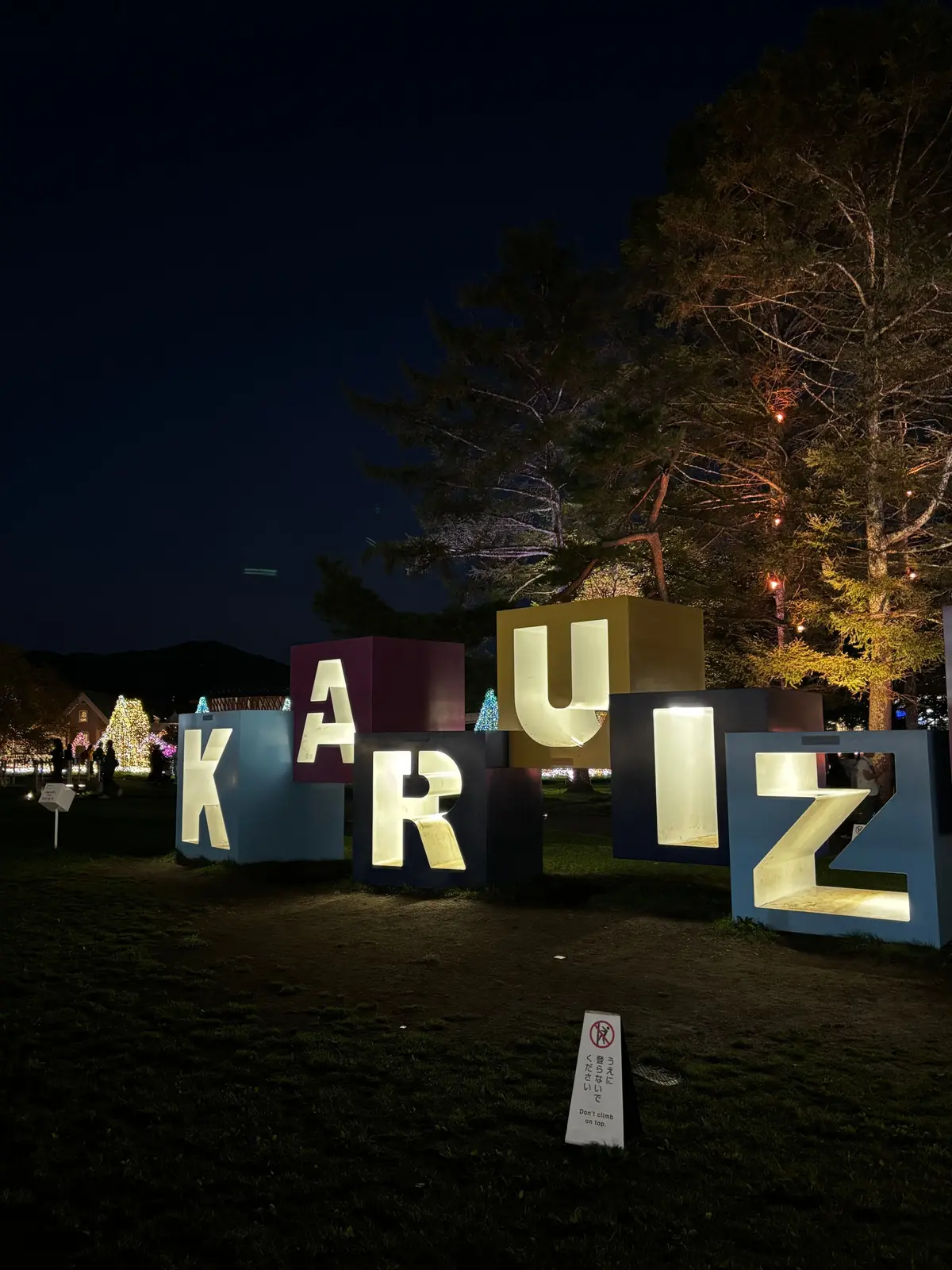 夜の公園に設置された、カラフルな大型のアルファベット「KA RUIZAWA」のオブジェ。背景には木々とライトアップされた建物が見える。