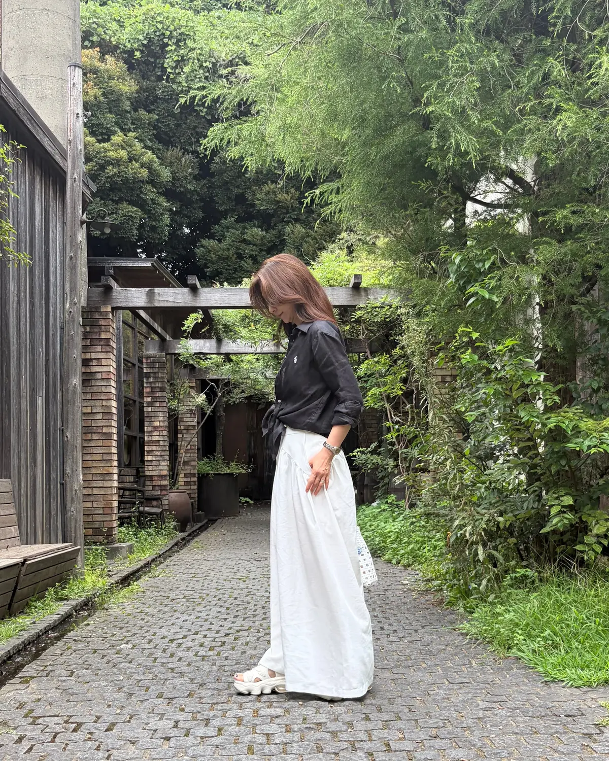 白いワイドパンツに黒シャツを合わせた女性が、石畳の小道に立っている全身写真。背景には緑豊かな木々と建物の壁が見える。