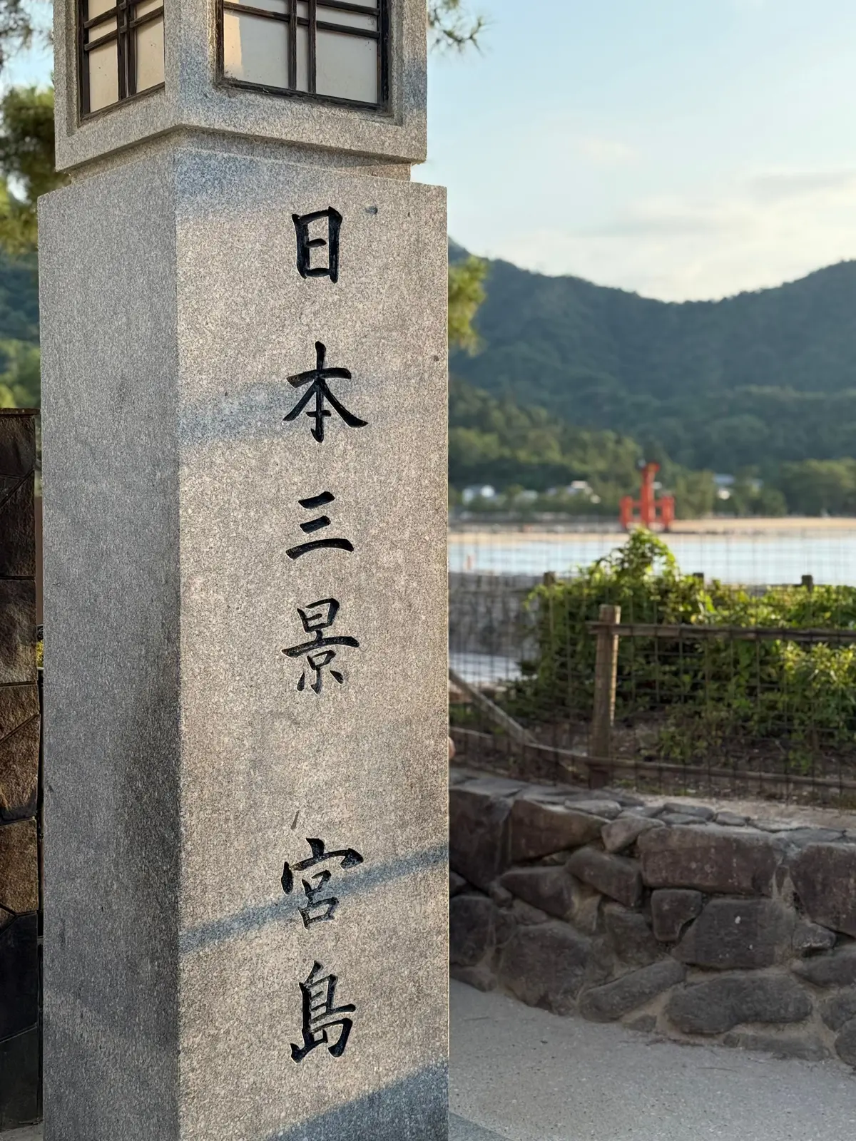 灰色の石柱に黒い文字で「日本三重県宮島」と刻まれ、背景に緑の山々と瀬戸内海の穏やかな水面が広がる風景。