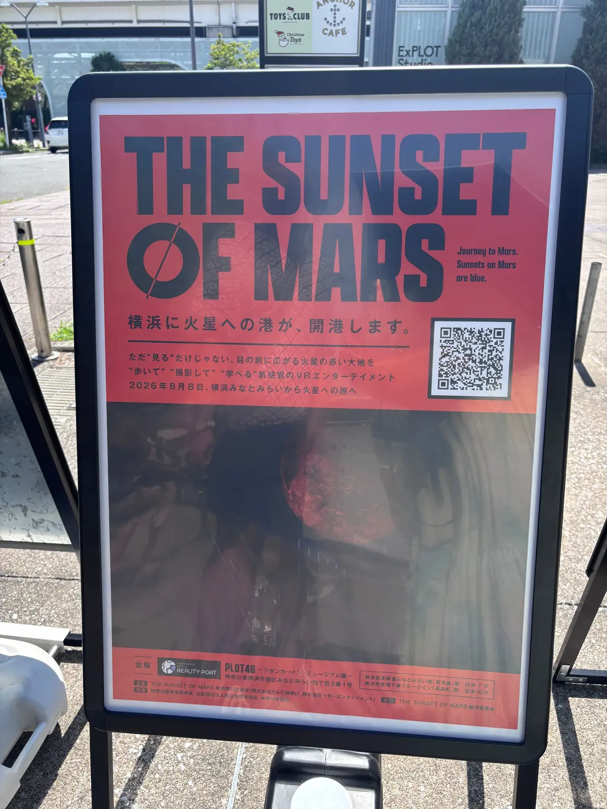街頭に設置された「THE SUNSET OF MARS」と題されたポスター。
夕焼けのイメージ写真が中央に大きく配置され、右上にQRコード、右下にはイベントの詳細情報が記載されている。背景には、店舗らしき建物が見える。