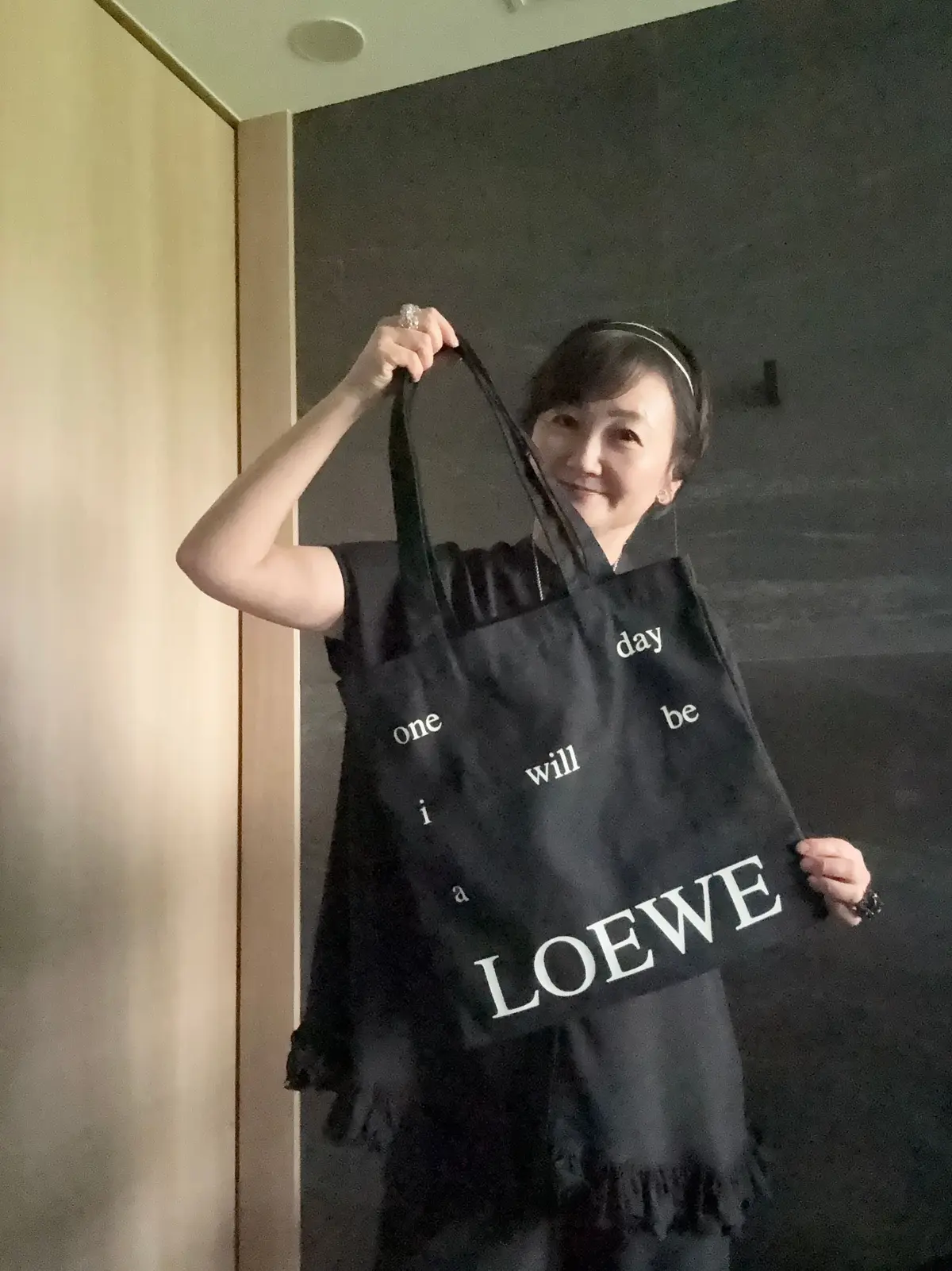 女性が黒いトートバッグを手に持つ。バッグには「LOEWE」というブランド名と「one day i will be」というフレーズが書かれている。背景は暗い壁。