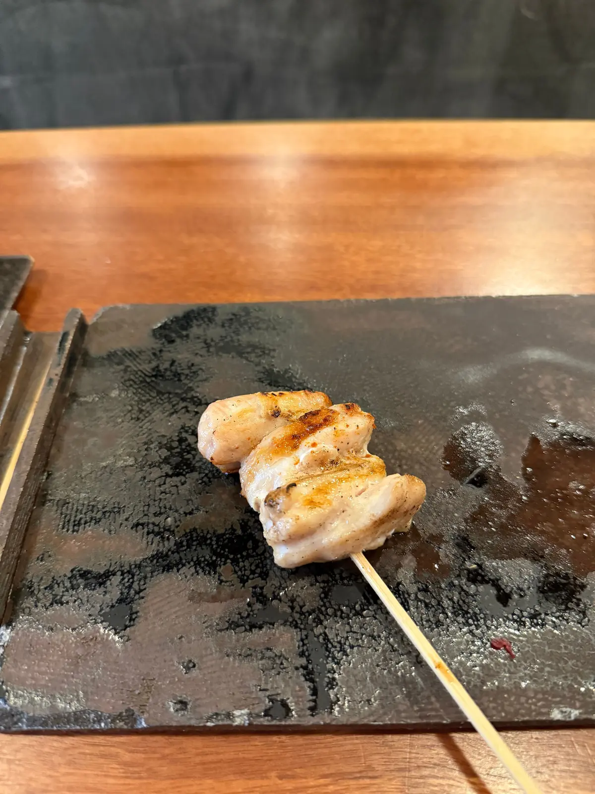 串に刺さった鶏肉の焼いたもの。表面は軽く焦げ付き、照りがある。黒い石板の上に置かれ、木製のテーブルに配置されている。