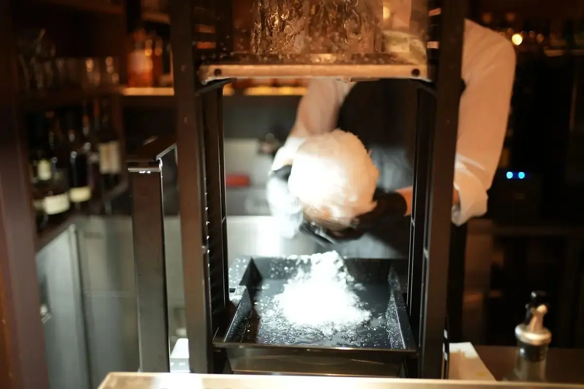 バーカウンターで、白いコックコートを着た人が氷を削っている様子。砕氷機の中には氷が詰まっており、背景には棚にグラスやボトルが並んでいる。