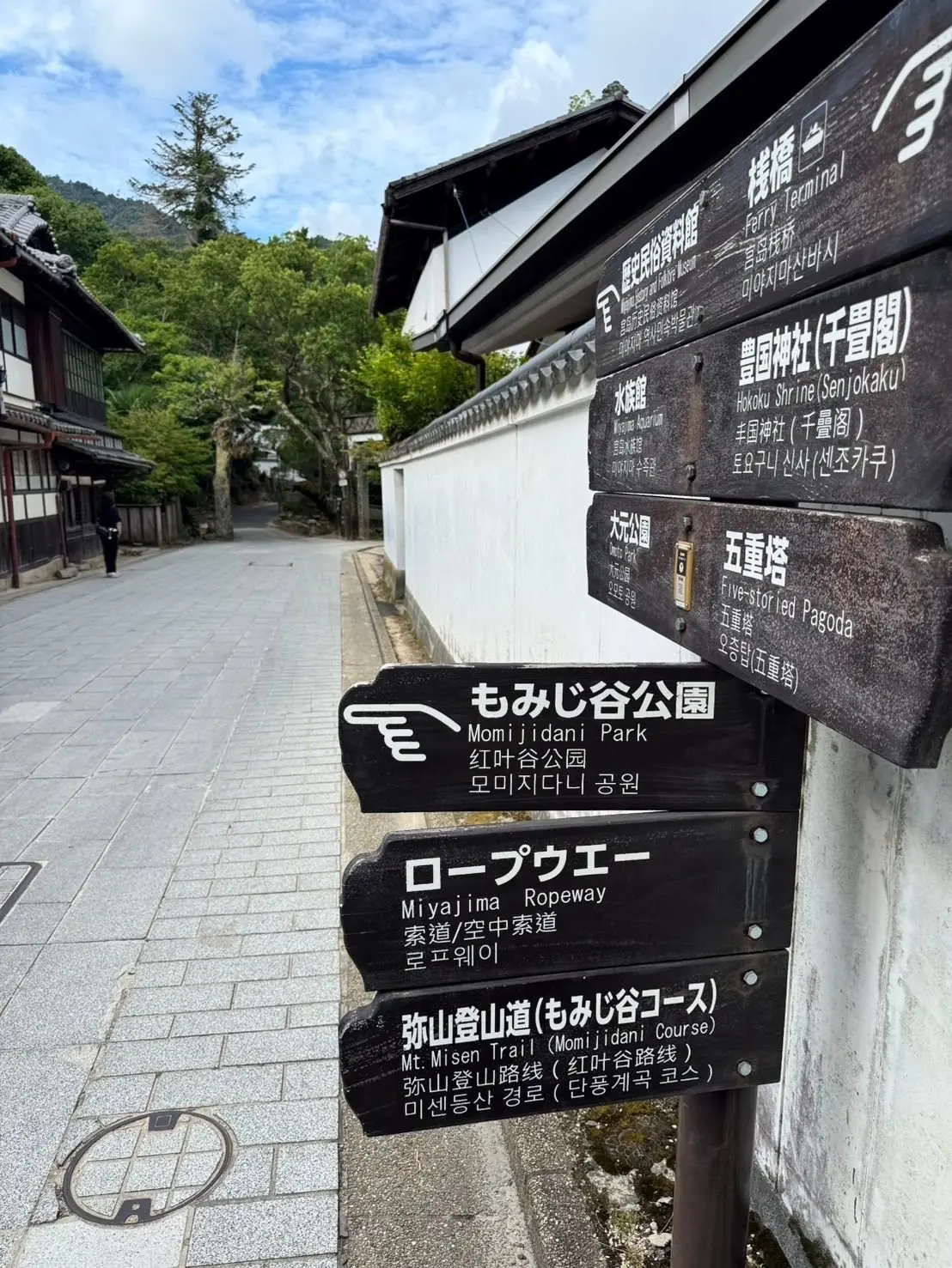 宮島の案内板。木製の多言語標識で、Momiiji Park、ロープウェイ、Mt. Misen Trailなどの観光スポットが記載されている。背景には、伝統的な木造建築と緑豊かな風景が見える。
