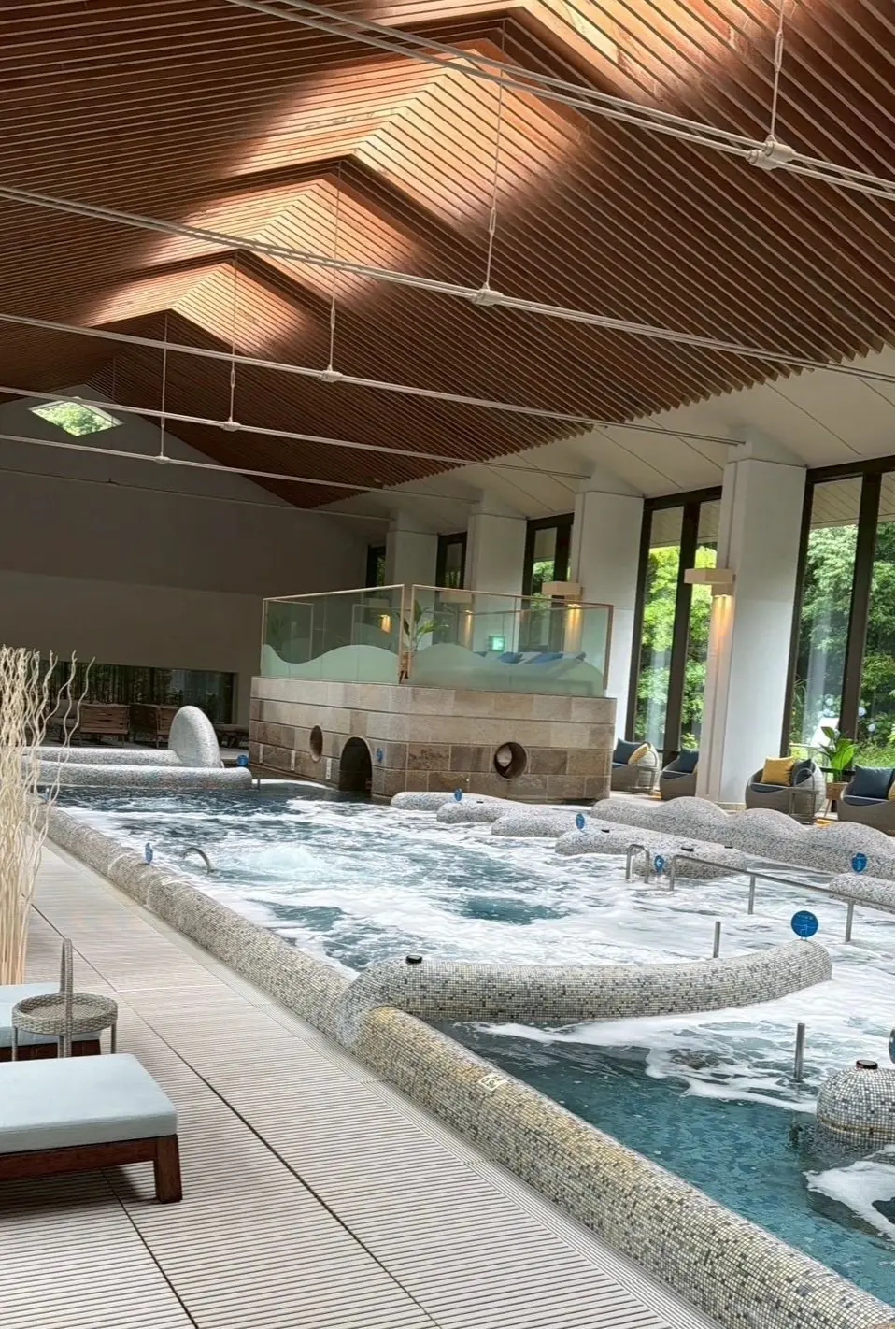 木製の天井と大きな窓を持つスパの屋内プールエリア。石造りのプールに泡が立ち、周囲にはリラックス用の椅子やソファが配置されている。