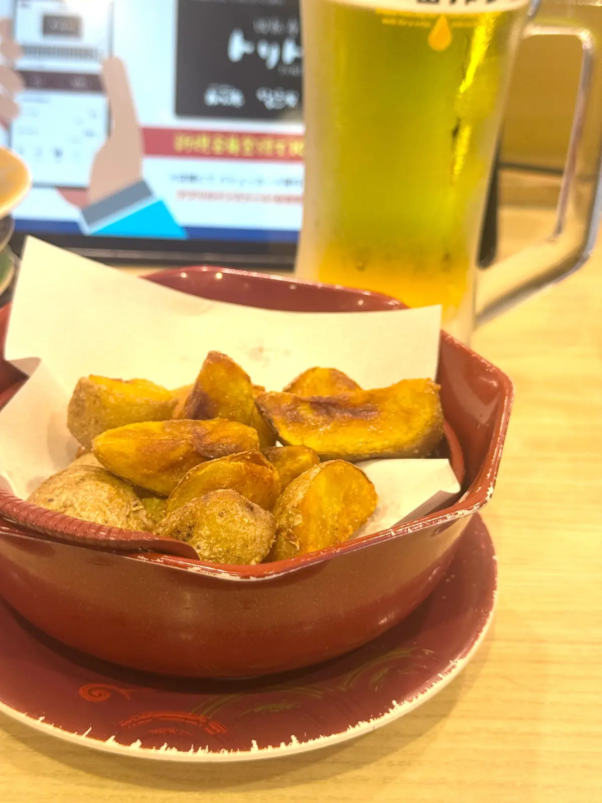 赤色の小皿に盛り付けられた、油で揚げられた様子のじゃがいも。その隣には、グラスに入った淡い色の飲み物。背景にはメニューらしきものが置かれている。