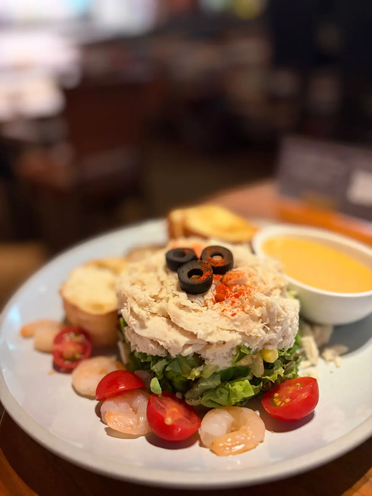 グレーの皿に盛り付けられた、鶏肉とレタスの層状サラダ。周りにはエビ、トマト、オリーブ、バゲット、ディップソースが添えられている。