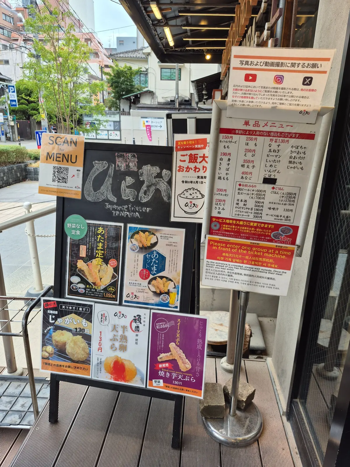映画館のチケット売り場の案内板。メニューや割引情報、注意事項などが記載され、チケットを購入した人が手すりにもたれている。背景には映画館のエントランスがある。