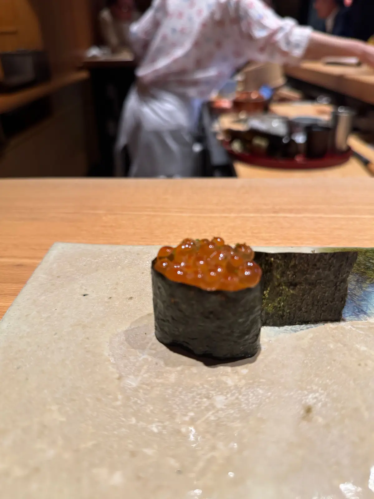 木のカウンターに置かれた、イカの握り寿司。表面にはオレンジ色のイクラが盛り付けられている。背景には厨房スタッフの姿が見える。