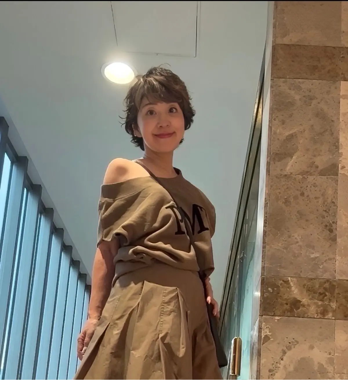 茶色のオフショルダーシャツとスカートを着た女性が、建物の階段で微笑んでいる。短い茶色の髪、やや俯瞰のアングル。背景には窓とタイル張りの壁。
