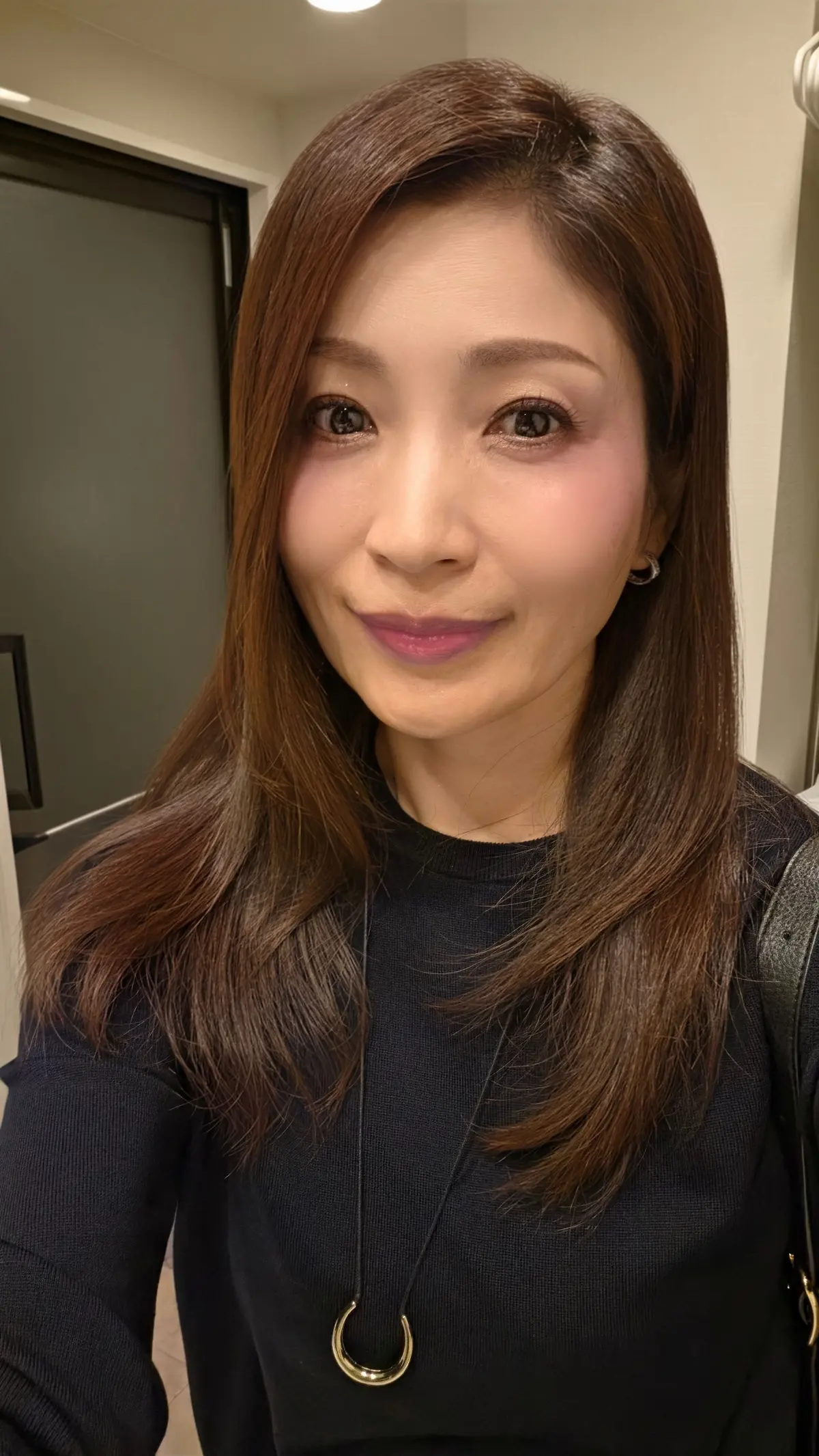 黒いカットソーを着た女性のポートレート。茶色のボブヘアで、ピンク系のリップを付けている。首には金色のC字型のネックレス。背景は白っぽい壁。