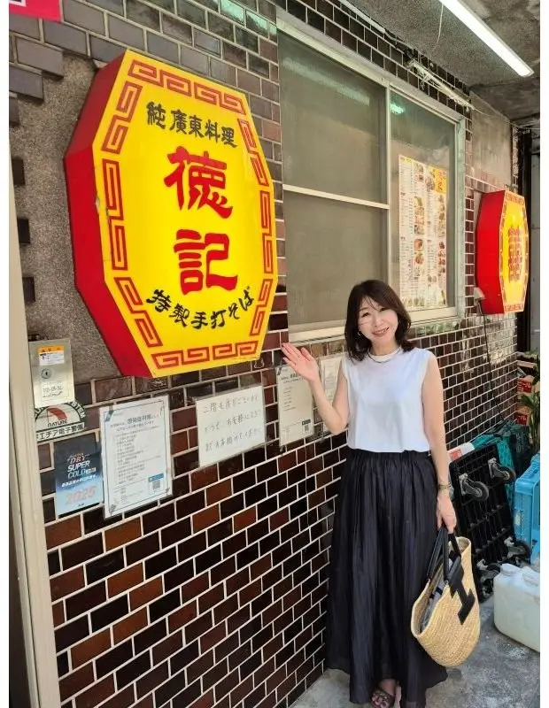 茶色のタイル張りの壁に、黄色の看板とメニューが貼られた店先。女性が笑顔でポーズを取り、右手にストローハットを持っている。