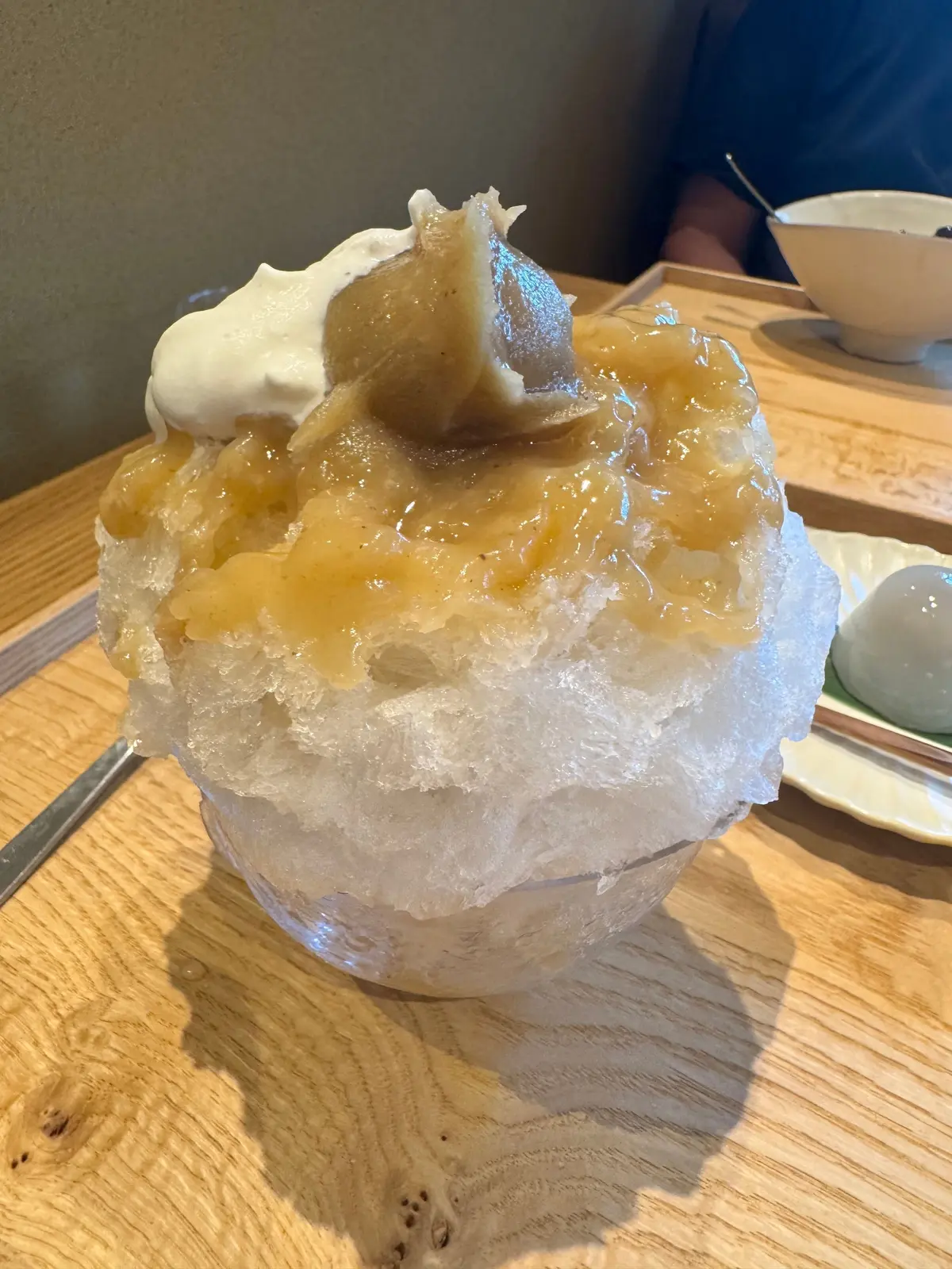 透明な器に盛り付けられた、細かく削られた雪氷の上に、蜜煮したような明るいオレンジ色の果肉と白いクリームが乗ったデザート。木製のテーブルに置かれ、背景には白い器と人物がぼやけて写っている。