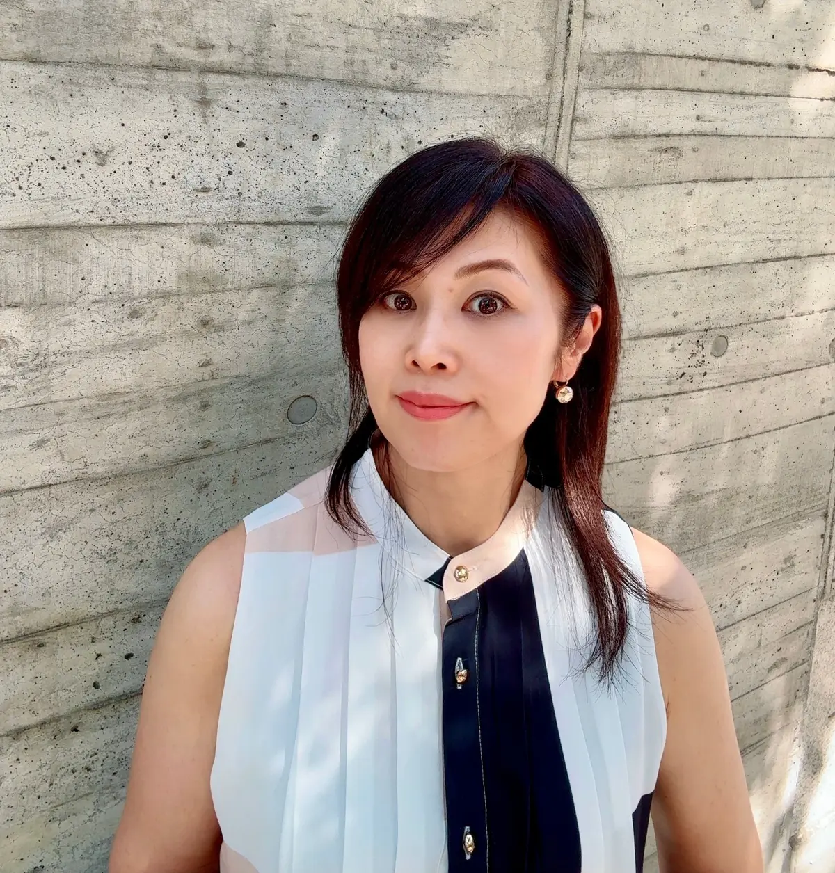 黒と白の装飾的なトップスを着た女性が、コンクリートの壁を背景にこちらを見ているポートレート。黒髪のボブカットで、控えめなアクセサリーを身につけている。