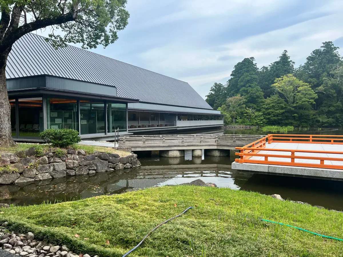 水辺に面したせんぐう館とオレンジ色の橋が特徴的な風景。建物はグレーの屋根と黒いフレームの大きな窓を持つ。手前に石積みと緑の芝生、水面には建物や橋の反射が見える。背景には緑豊かな木々が茂る。