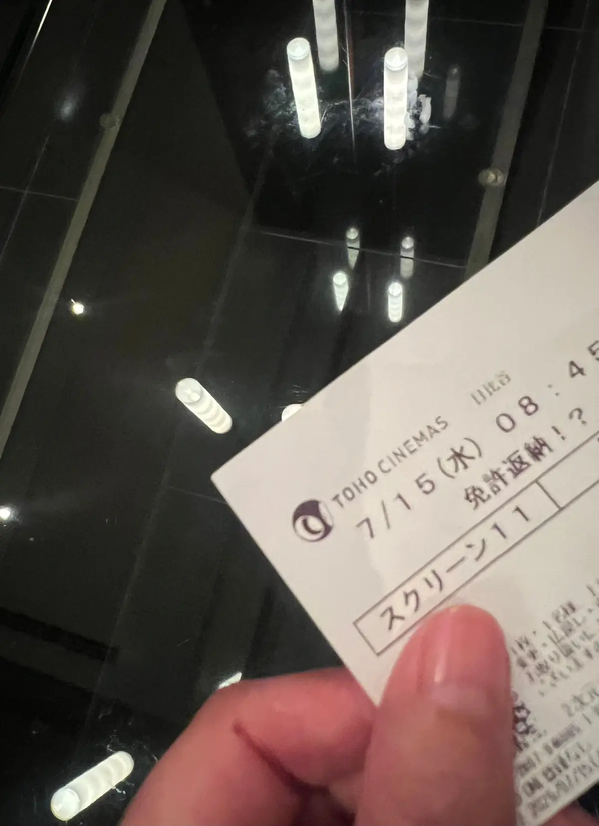 映画館の床に置かれた、日本の映画館チェーンTOHO CINEMASの入場券。7月15日午前8時45分の上映、スクリーンの11番を記した白い紙切れ。背景には、床に反射する照明の光が縦に並んでいる。