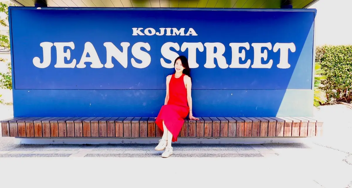 鮮やかな赤いワンピースを着た女性が、木製のベンチに座ってこちらを見ている。背景には青色の「KOJIMA JEANS STREET」の文字が描かれた壁がある。