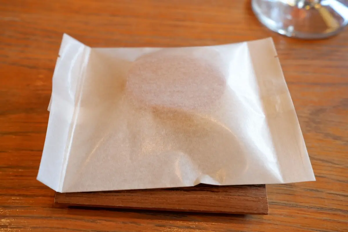 木製の台の上に置かれた、和紙のような包み紙で包まれた丸いお菓子。表面には淡いピンク色の粉がふられている。背景にはグラスが見える。