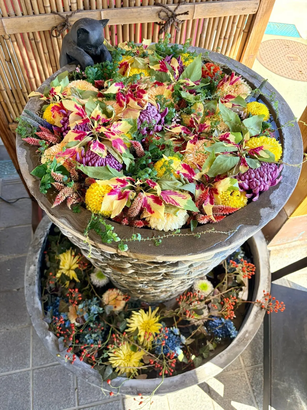 石造りのプランターに、赤や黄色の花、紫の穂状の花、緑の葉が鮮やかに配置されたアレンジメント。プランターの下段には、同様の花々が散りばめられている。背景には竹製の柵が見える。