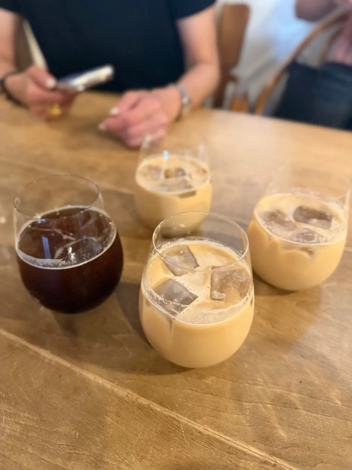 木製テーブルに並んだ4つのグラス、左から濃いコーヒー、ミルクティー、アイスコーヒー、ミルクティー、氷とストロー入り