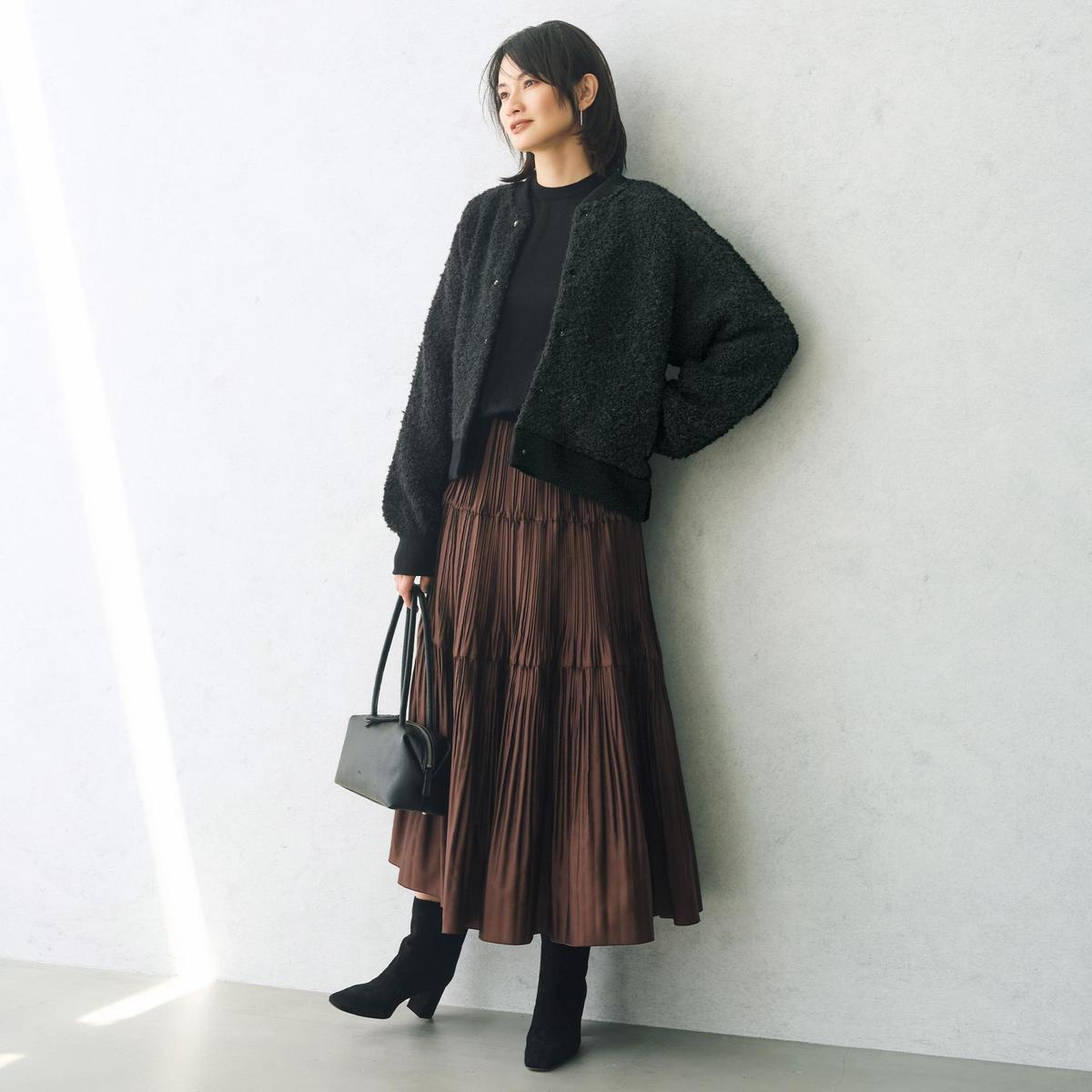 黒いカットソーの上に黒のモヘアカーディガン、深みのある茶色のプリーツスカートを合わせた女性。黒いニーハイブーツと黒いハンドバッグを身につけ、白い壁の前に立っている全身像。