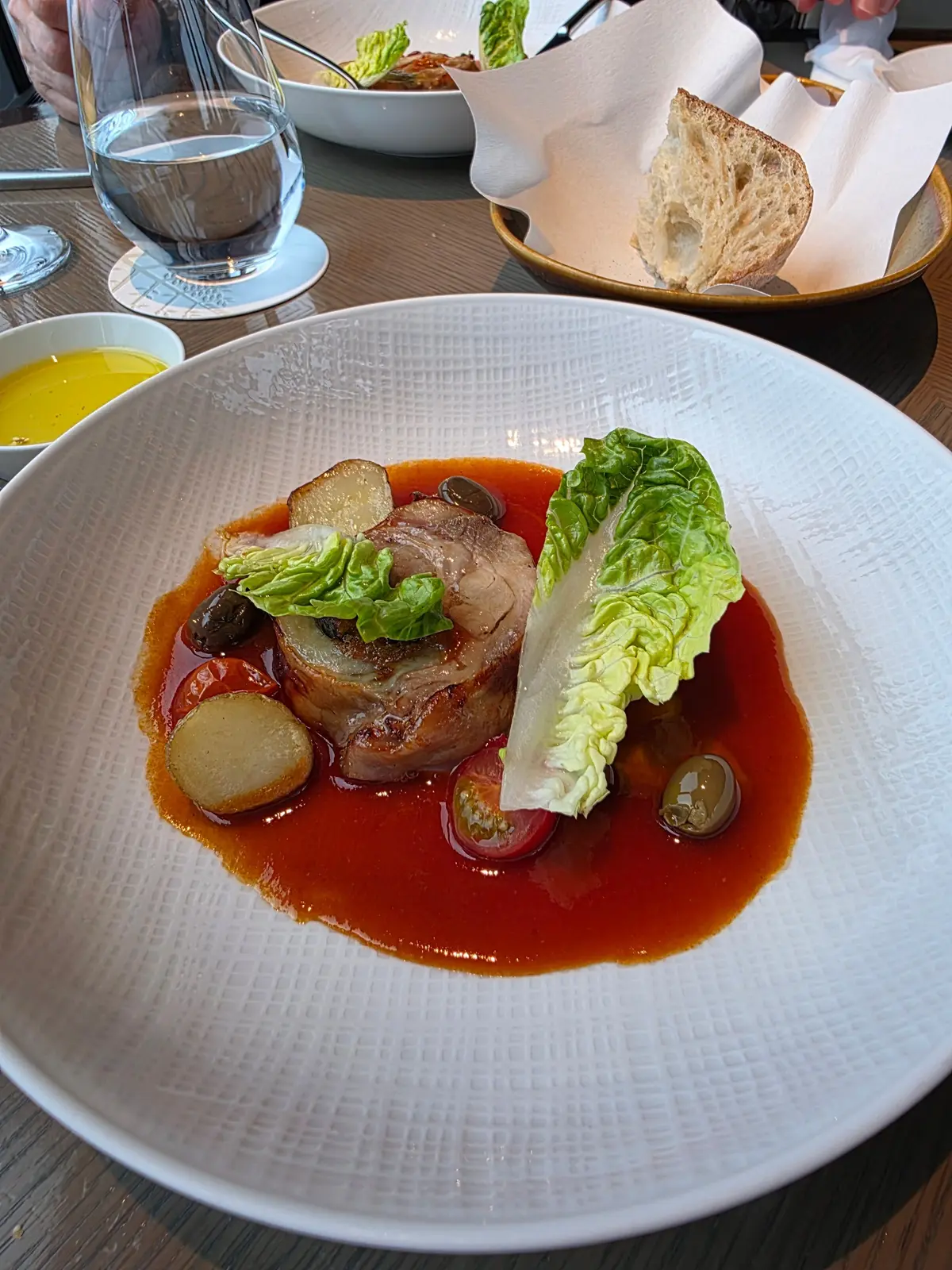 白磁の皿に盛り付けられた、赤色のソースが広がる料理。中心には肉が置かれ、その周りにレタス、トマト、オクラ、ピクルスなどの具材が彩られている。右にはパン、左には水が置かれている。
