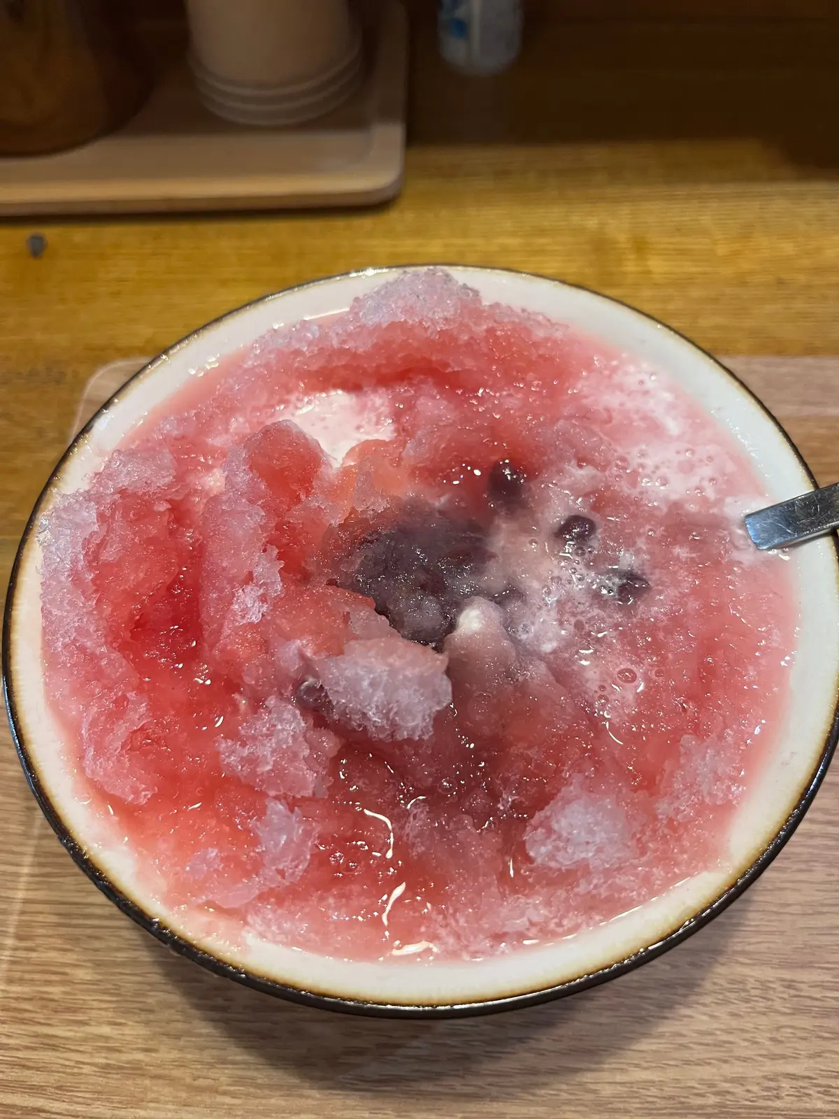 ピンク色のシロップが染み込んだかき氷が、茶色の縁の白い器に入っている。器の奥には木製のテーブルと、白いカップが置かれた木製のトレーが見える。