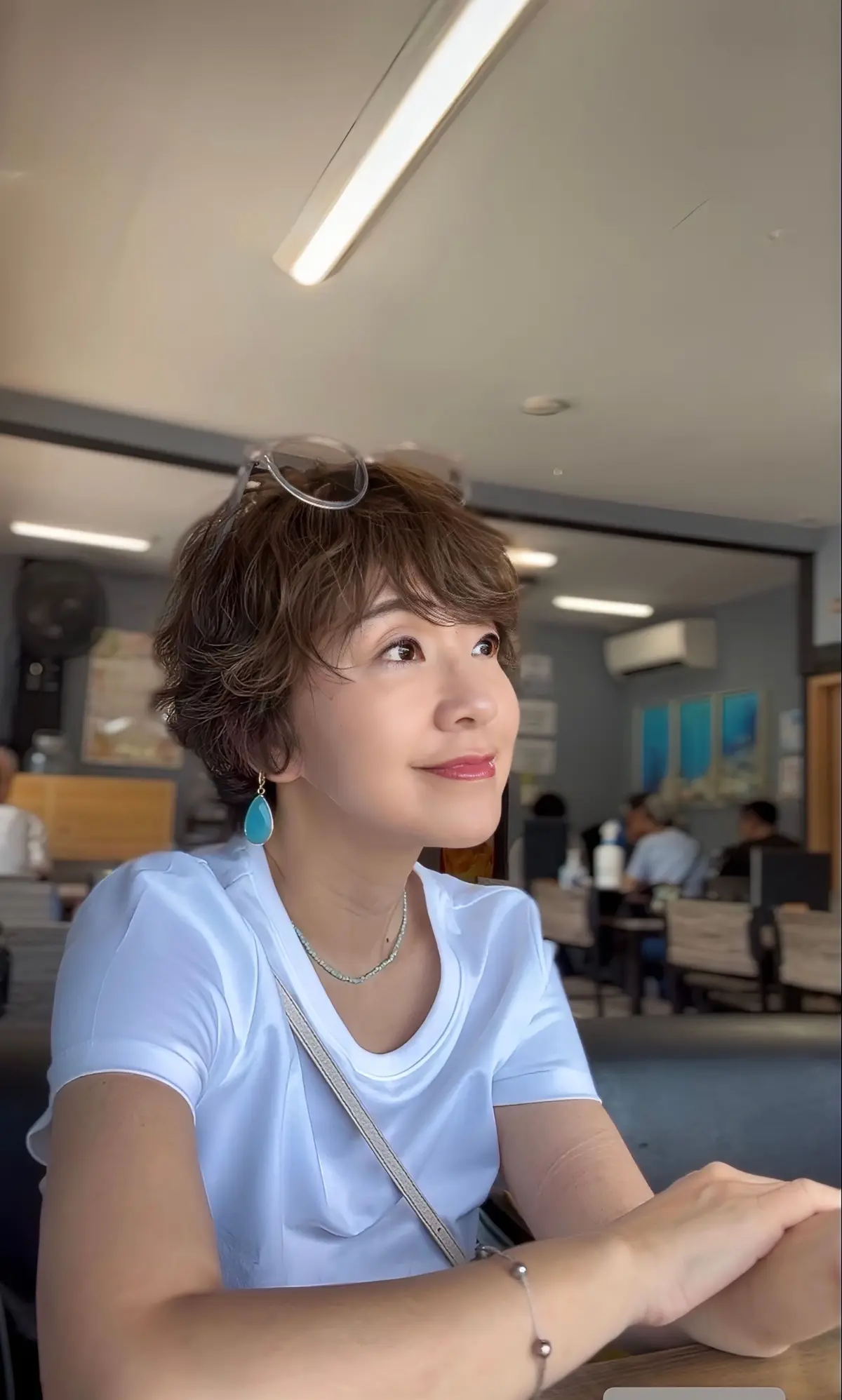 白いTシャツを着た女性が、カフェで遠くを見上げている。茶色の短い髪に、水色のイヤリングとネックレスを身につけている。背景には、テーブル席と窓が見える。