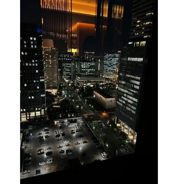 夜の都市の俯瞰。高層ビル群が立ち並び、窓に光が灯る。手前には駐車場と道路が広がり、車のライトが点滅している。オレンジ色の光が建物の隙間から差し込んでいる。