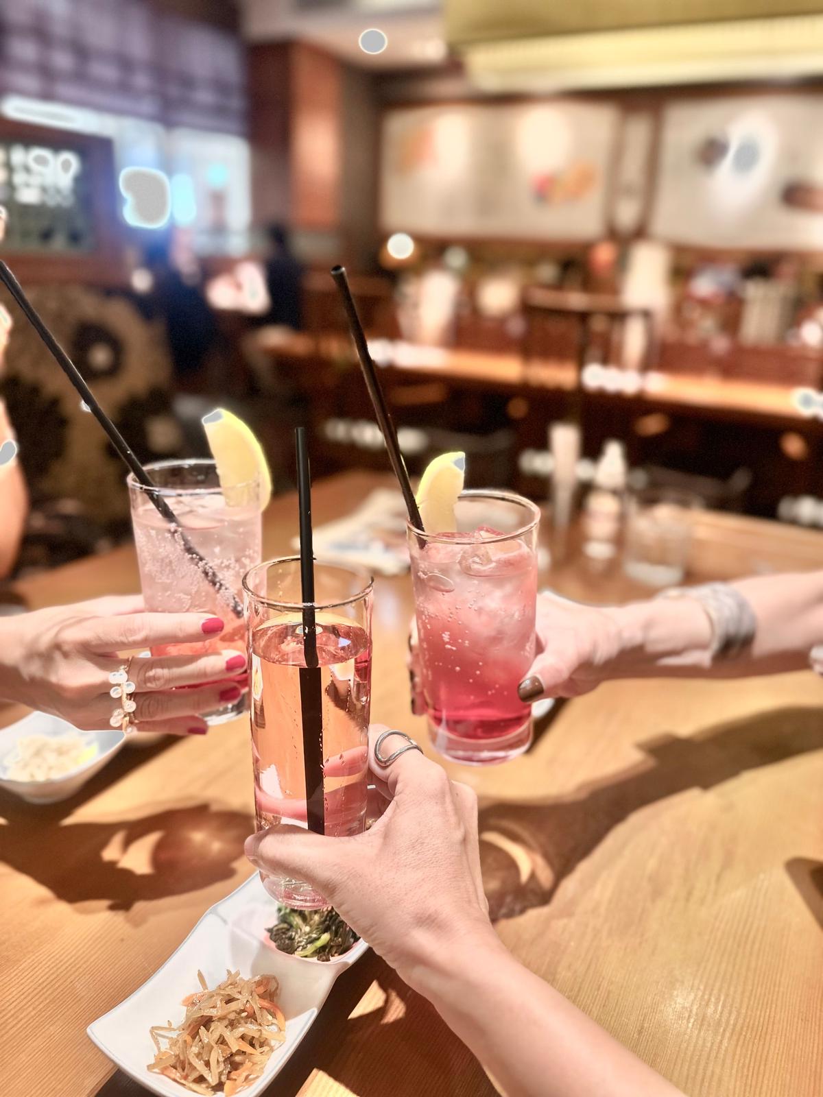 木製のテーブルに置かれた3つのグラスが、手で持ち上げられて乾杯している様子。ピンクや透明の飲み物にレモンのスライスが添えられ、黒いストローが挿さっている。手前には小皿に料理が盛り付けられている。背景にはレストランのぼんやりとした様子が見える。