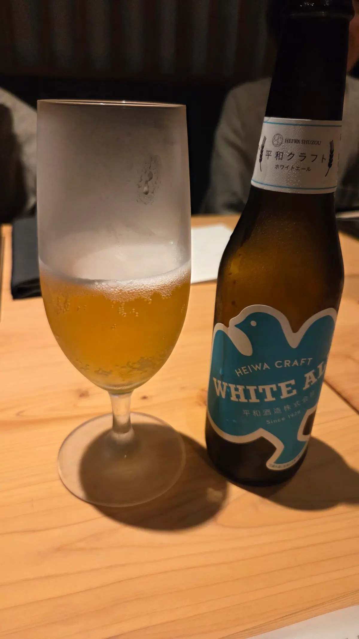 木製のテーブルに置かれた、平和クラフトホワイトエールビール。グラスには泡立ち、瓶には青いラベルが貼られている。背景には人物がぼやけて写っている。