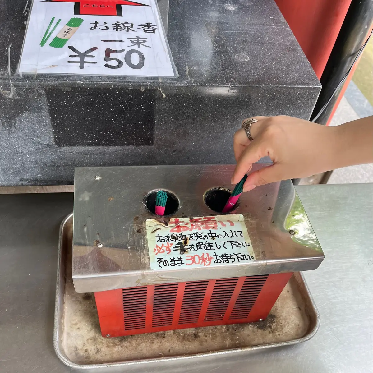 お茶の自動販売機。緑色のパックお茶が並び、価格は1本50円。投入口に手が入っており、説明書きが貼られている。
