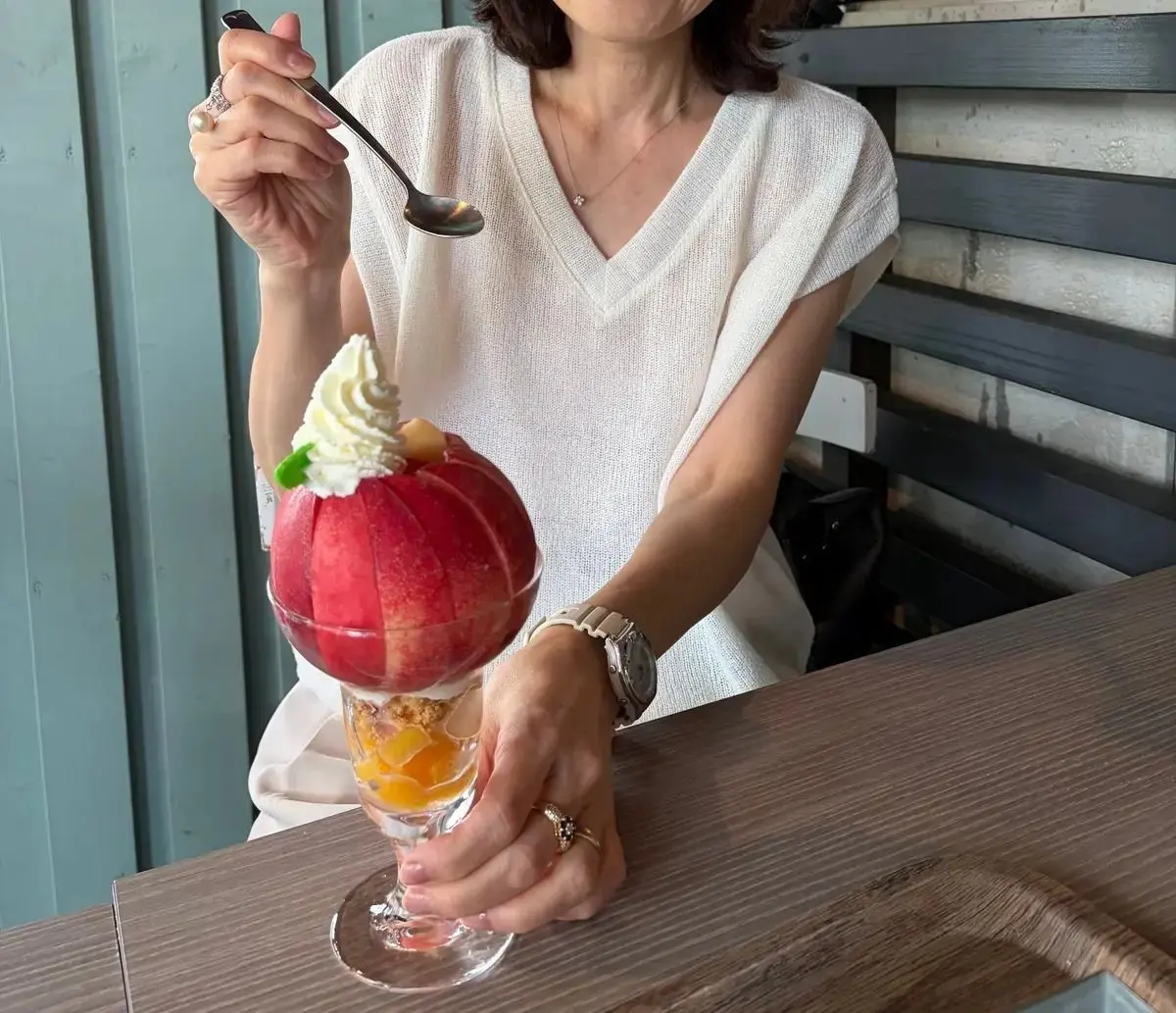白いVネックのトップスを着た女性が、ガラスの器に入ったイチゴのかき氷を手に持っている。かき氷は丸く盛り付けられ、ホイップクリームとフルーツが添えられている。女性はスプーンを持ち、かき氷を食べる様子。
