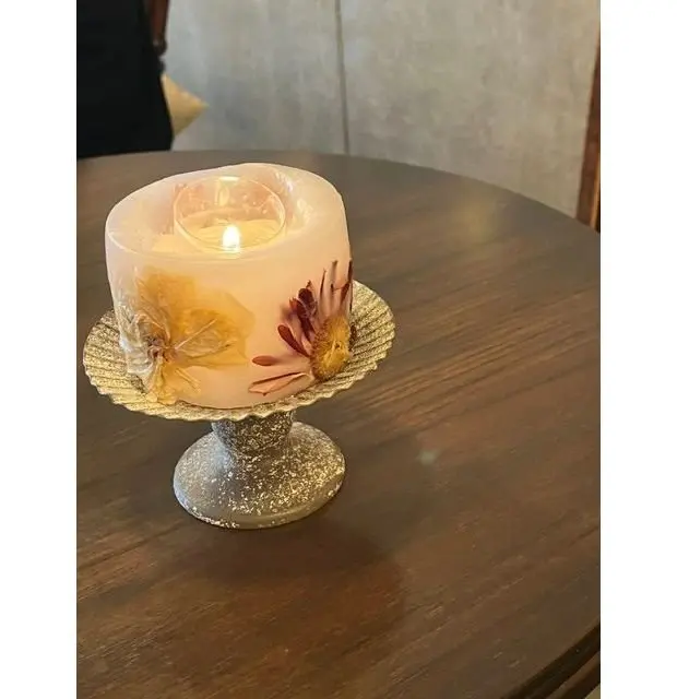 木製のテーブルに置かれた、花びらが埋め込まれたキャンドルと、銀色のペーパーナプキンホルダー。キャンドルは点灯しており、温かい光を放っている。