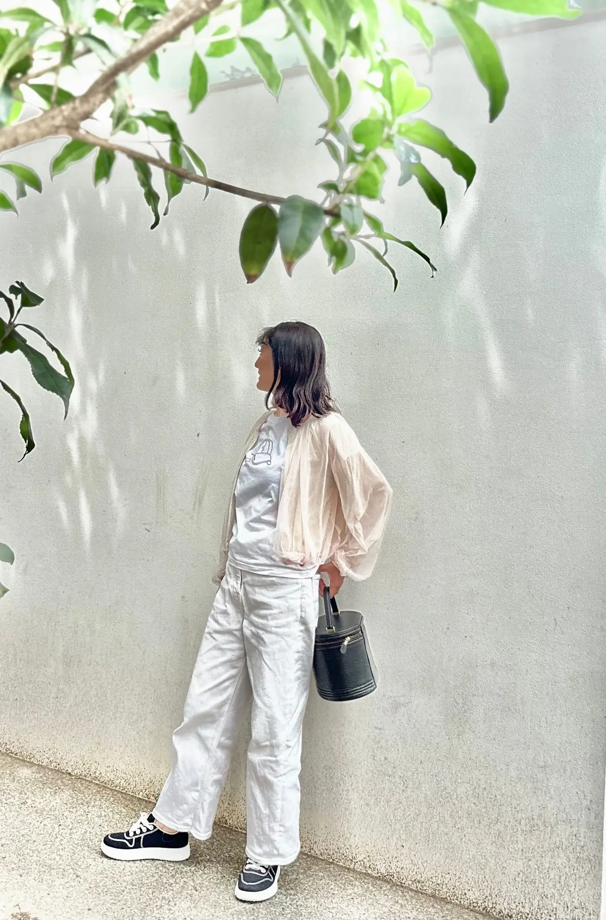 女性が白い壁に寄りかかり、左を向いて立っている。薄手のベージュのショール、シルバーのTシャツ、ゆったりとした白いズボン、黒いスニーカーを着用。左手には小さな黒いハンドバッグを下げている。背景には緑の葉が見える。