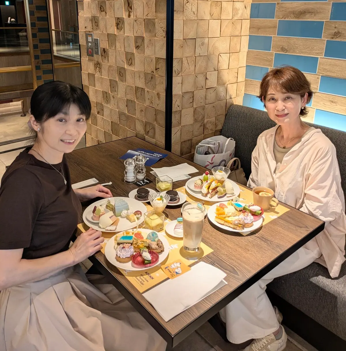 木製のテーブルに多彩なデザートと飲み物が並び、2人の女性が笑顔で座っている。壁は木製と青いタイルで装飾されている。