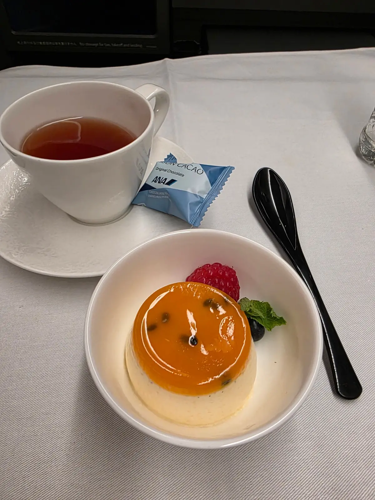 白いテーブルに置かれた、ANAの機内食。黄色のプリン、赤色のラズベリー、緑色のミントが添えられている。カップに入ったお茶と、カカオのチョコレートも写っている。