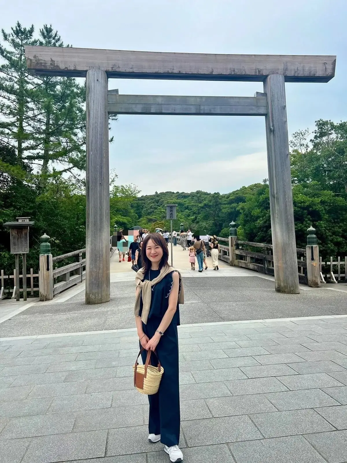 木造の鳥居の前で、黒いタンクトップとワイドパンツを着た女性が微笑んでいる。右手に茶色のバッグを持ち、肩には黄色いスカーフをかけている。背景には緑豊かな木々と、歩行者がいる石畳の道が見える。