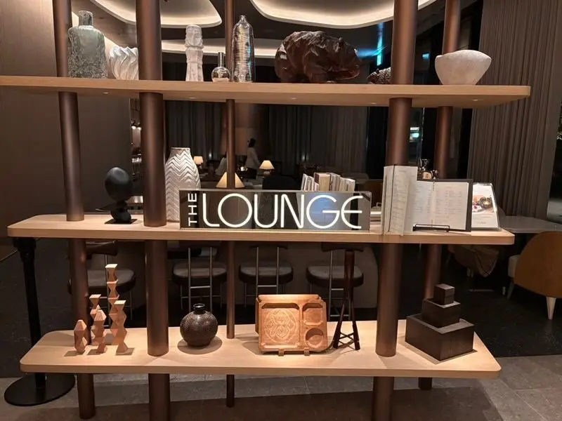 木製の棚に、様々な装飾品やメニューが置かれている様子。中央にはネオンサインで「THE LOUNGE」と表示されている。背景には、落ち着いた色のソファや椅子が並んだラウンジの様子が見える。