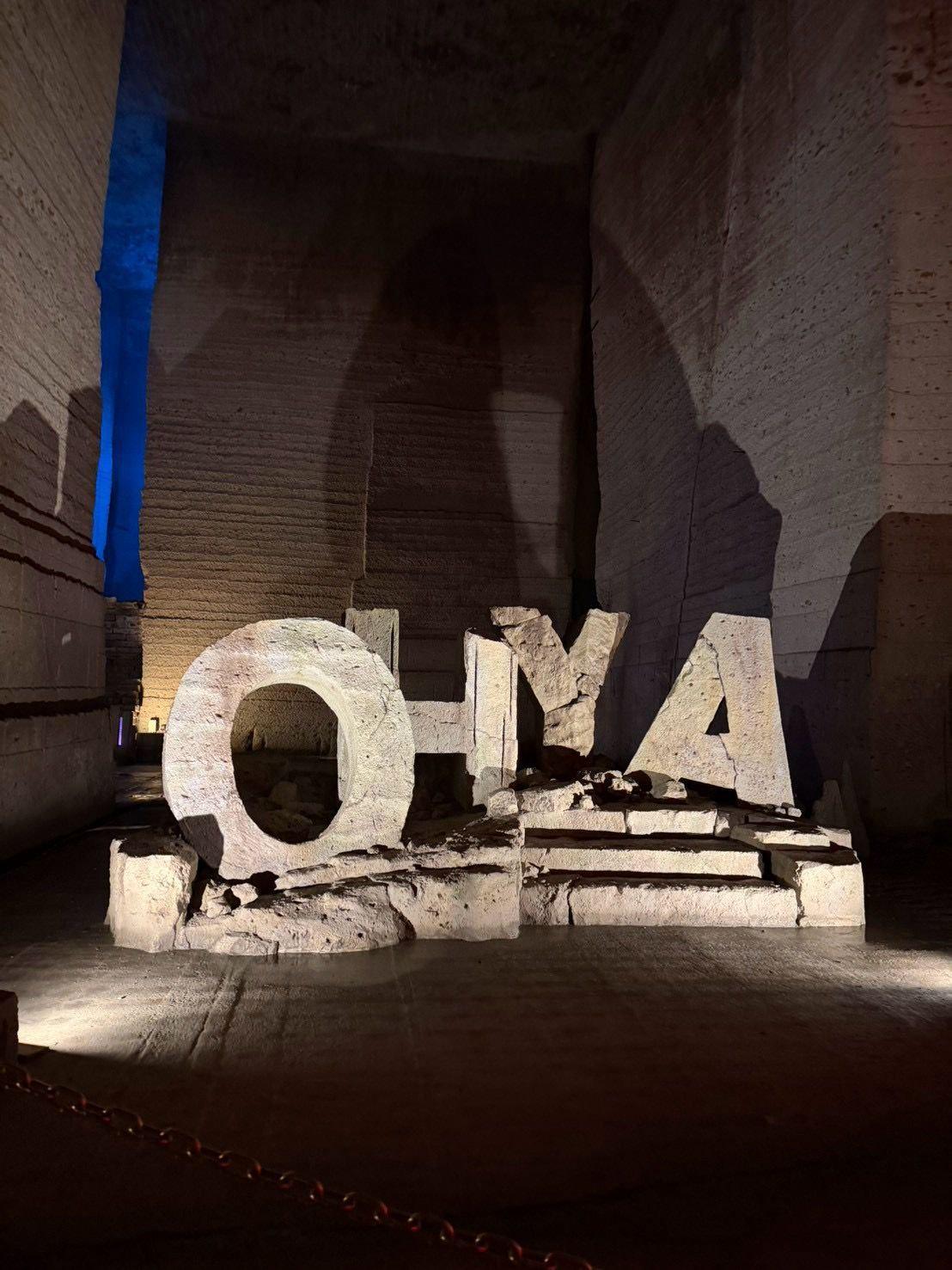石造りの壁に囲まれた空間に、白い大きな文字「OYA」が置かれている。文字は積み石のように作られており、床には赤いロープが設置されている。背景には青いライトアップが見られる。