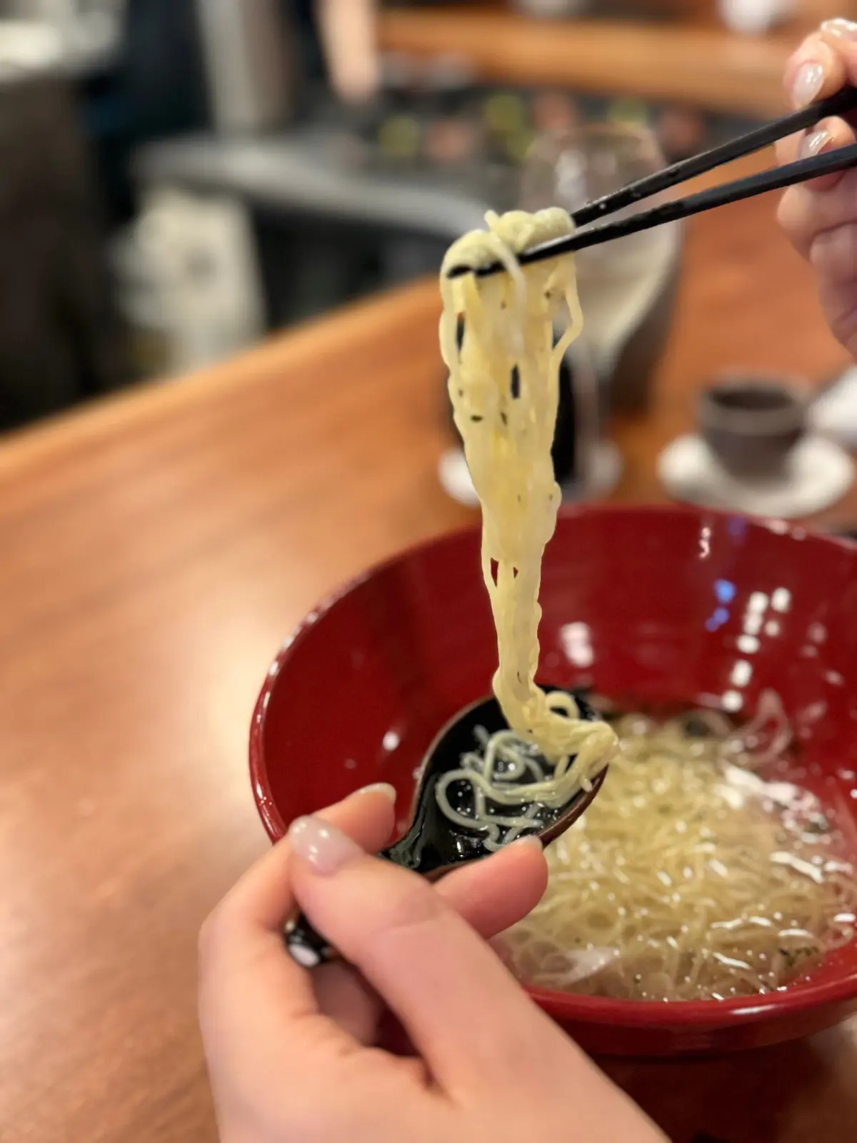 大阪・福島の人気焼き鳥店「焼き鳥 小松」でいただいた焼き鳥コース。赤いボウルに入った〆の冷麺を、はしとスプーンで掴んでいる様子。麺は黄色く、スープの中に浮かんでいる。さっぱりレモン風味